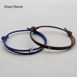 May include: Two adjustable bracelets. One is navy blue with a star-shaped charm, the other is brown with an X-shaped charm. Both bracelets have a knotted closure. The brand name "Grass Horse" is in the top left corner.