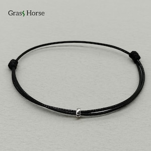 Puede incluir: Pulsera de cordón negro con una pequeña cuenta de color plateado. La pulsera está anudada en ambos extremos. La marca "Grass Horse" está en la esquina superior izquierda.