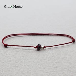 May include: A minimalist red string bracelet featuring a dark red bead and a small silver bead. The bracelet is simple and elegant, suitable for everyday wear. The brand name "Grass Horse" is in the top left corner.