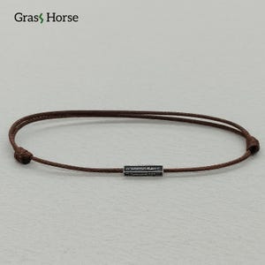 Könnte beinhalten: Ein braunes Kordelarmband mit einem kleinen, rechteckigen, silberfarbenen Akzent. Das Armband ist schlicht und minimalistisch und eignet sich für den täglichen Gebrauch. Der Markenname "Grass Horse" befindet sich oben links.