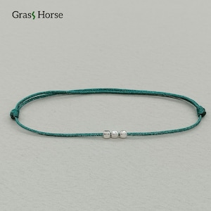 Può includere: Un braccialetto color teal con tre piccole perline d'argento. Il braccialetto è realizzato con un cordoncino sottile e intrecciato. Il marchio "Gras Horse" è visibile in alto a sinistra.
