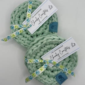 May include: Two light green, chunky knit cotton coasters. Each coaster is round and tied with a ribbon featuring peace signs. The coasters have tags that read "I'M HANDMADE Chunky Coasters 100% COTTON".