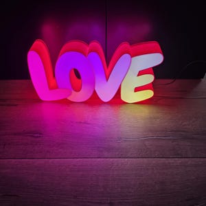 Peut inclure: Enseigne au néon avec le mot "LOVE". Les lettres sont bordées de rouge avec un contour blanc et remplies de lumière rose, violette et jaune. L'enseigne est posée sur une surface en bois.