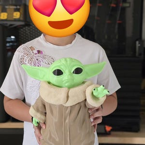 Peut inclure: Une figurine en plastique vert représentant une petite créature aux grandes oreilles pointues et aux yeux noirs, vêtue d'un manteau beige avec un col et des poignets épais en tissu texturé crème. La figurine a une finition lisse.