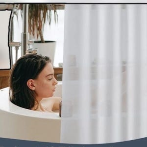 May include: A woman relaxing in a white bathtub. The bathroom features a white shower curtain and a silver faucet. A potted plant is visible in the background, adding a touch of nature to the scene. The overall aesthetic is clean and minimalist.