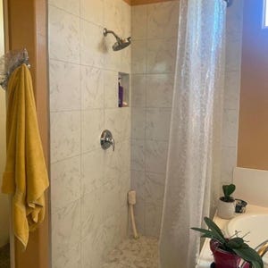 May include: A bathroom shower with white marble-look tiles, a silver shower head, and a clear shower curtain. A yellow towel hangs on a hook. A small niche holds a purple bottle. A plant sits on the edge of a bathtub.