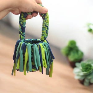 May include: A small green and blue crocheted basket with a handle. The basket is made from strips of fabric and has a fringe around the bottom.