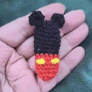 May include: A small, handmade crochet Mickey Mouse figure. The figure features black ears and head, a red body with yellow accents, and is held in a person's hand. The craft item is likely a keychain or small toy.