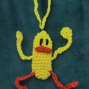 May include: A yellow crochet figure with black eyes, a red mouth, and red legs. The figure has raised arms and a loop at the top for hanging. The background is a dark green fabric.