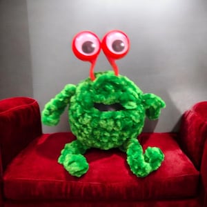 Può includere: Un peluche verde a forma di mostro con grandi occhi mobili su steli rossi. Il giocattolo morbido e strutturato ha un corpo rotondo, braccia e gambe ed è seduto su un divano rosso. Il mostro ha una bocca ampia e aperta.