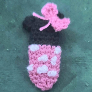 Pode incluir: Um item de crochê feito à mão em forma de cabeça de personagem de desenho animado. A parte superior é preta, com um laço rosa. O corpo é rosa com detalhes brancos em forma de nuvem. Este item é provavelmente um acessório decorativo ou um brinquedo.