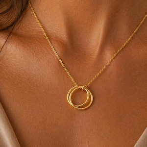 May include: A gold-colored necklace featuring two interlocking circles as a pendant. The delicate chain and pendant are displayed against a skin tone background, highlighting the jewelry's design and color. The necklace is a simple yet elegant accessory.