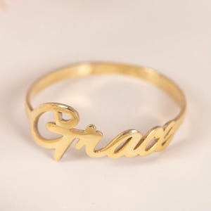 Könnte beinhalten: Ein goldfarbener Ring mit dem Namen "Grace" in Schreibschrift. Der Ring liegt auf einem weichen, cremefarbenen Hintergrund, mit einigen getrockneten Blumen in der oberen linken Ecke. Ein zarter Schmuck.