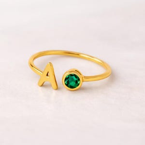 Puede incluir: Anillo dorado con la letra "A" y una gema redonda de color verde esmeralda. El anillo tiene un diseño abierto. La gema tiene aproximadamente 0,6 cm de diámetro.