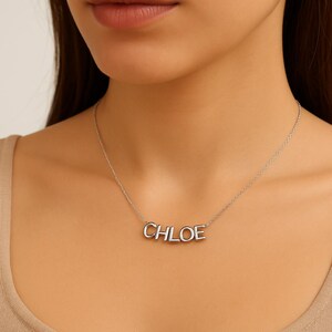 May include: A silver necklace spelling "CHLOE" in capital letters. The necklace is worn around the neck. The chain is fine and the letters are in a block style. The necklace is a simple, elegant piece.