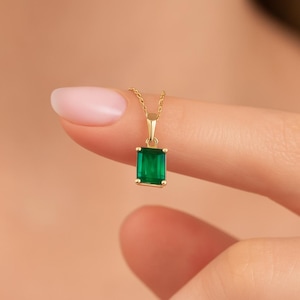 Op de afbeelding: Een gouden ketting met een rechthoekige smaragd hanger. De smaragd is heldergroen en is gezet in een gouden bezel. De ketting is delicaat en lijkt van goud te zijn. De achtergrond is zacht perzik van kleur.