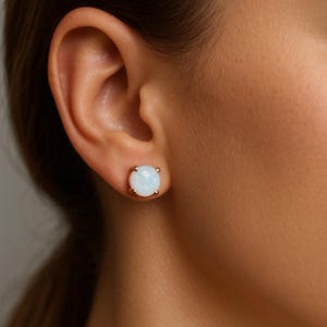 May include: A close-up of a person's ear with a gold stud earring. The earring features a round, light blue opal stone set in a gold bezel. The earring is a classic stud design.