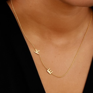 Może przedstawiać: Delikatny złoty naszyjnik z literami "K" i "E". Litery wisiorka połączone są cienkim łańcuszkiem. Naszyjnik jest noszony na ciemnym tle.