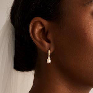 Könnte beinhalten: Nahaufnahme eines goldenen Creolen-Ohrrings mit einer weißen, tropfenförmigen Perle. Der Ohrring wird am Ohr einer Person getragen. Der Hintergrund ist unscharf, wodurch der Fokus auf dem Schmuck liegt.