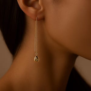 May include: A close-up of a gold earring with a chain and a teardrop-shaped, olive-green gemstone. The earring is dangling from the ear. The background is blurred.