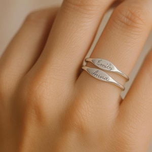 May include: Two silver rings with an oval design, stacked on a finger. The top ring is engraved with the name "Emily", and the bottom ring has the name "Anna". The rings are displayed on a light skin tone.