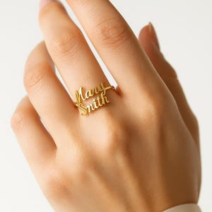 May include: A gold-colored ring with the names "Mary Smith" in a cursive font. The ring is on a finger, and the background is a soft, neutral color. The ring is a personalized jewelry item.