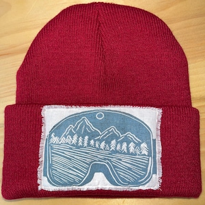 May include: A red knit beanie with a folded brim. The beanie features a white patch with a blue graphic of mountains, trees, and a sun or moon. The patch is rectangular with a frayed edge.