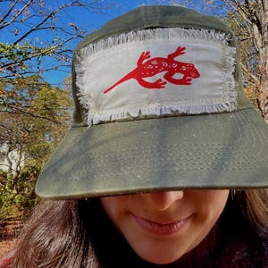 May include: Olive green bucket hat with a white patch featuring a red lizard design. The patch has a frayed edge. The hat is made of a durable fabric and has a wide brim, suitable for sun protection.