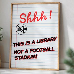 Op de afbeelding: Een ingelijste print met een houten frame. De print heeft een achtergrond van gelijnd papier met de rode tekst "Shhh!" bovenaan, een geluid-uit-pictogram en de tekst "THIS IS A LIBRARY NOT A FOOTBALL STADIUM!" in rood en zwart.