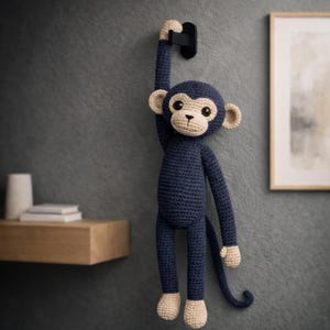 Könnte beinhalten: Ein marineblauer Häkelaffe hängt an einem schwarzen Haken an einer grauen Wand. Der Affe hat beige Akzente auf Gesicht, Ohren, Händen und Füßen. Der Schwanz des Affen ist eingerollt. Ein Holzregal und ein gerahmtes Kunstwerk befinden sich im Hintergrund.
