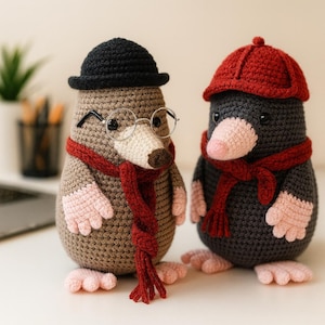 Puede incluir: Dos muñecos de topo hechos a mano con ganchillo. Uno es marrón con sombrero negro, gafas y bufanda roja. El otro es gris con gorra y bufanda rojas. Ambos tienen manos y pies rosas y están de pie.