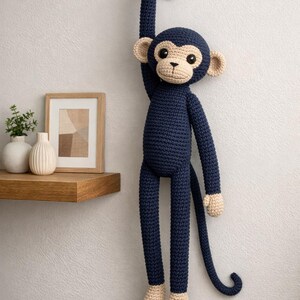 Può includere: Una scimmia all'uncinetto blu navy appesa a un gancio nero su un muro bianco. La scimmia ha accenti beige sul viso, sulle orecchie, sulle mani e sui piedi. Sono visibili gli arti lunghi e la coda della scimmia. Sullo sfondo una mensola in legno con oggetti decorativi.