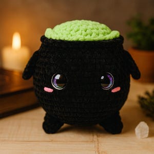 以下が含まれることがあります： 明るい緑色のトップ、大きく光沢のある目、ピンクの頬を持つ黒いかぎ針編みの大釜。大釜には小さなハンドルと足が付いています。背景には、ろうそくの火と小さな植物があり、気まぐれで手作りの美学を作り出しています。