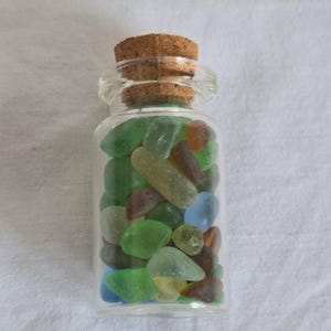 Könnte beinhalten: Eine kleine, klare Glasflasche gefüllt mit farbigem Meeresglas. Die Meeresglasstücke sind in verschiedenen Grüntönen, Blau, Braun und Orange gehalten. Die Flasche ist mit einem Korken verschlossen.