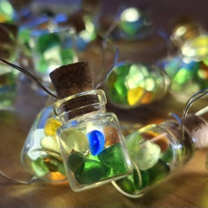 Op de afbeelding: Slinger van decoratieve lichtsnoeren met kleine glazen flesjes gevuld met kleurrijk zee glas. Elke fles heeft een kurk en bevat een klein LED-lampje, wat een warme gloed creëert. De stukjes zee glas zijn groen, blauw, geel en oranje.