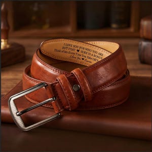 Op de afbeelding: Een opgerolde bruin lederen riem met een zilveren gesp. De binnenkant van de riem heeft gegraveerde tekst die luidt "I LOVE YOU FOR WHO YOU ARE BUT THAT SURE IS A BONUS Think of Me Every Time You Take Off I Love You Lauren."