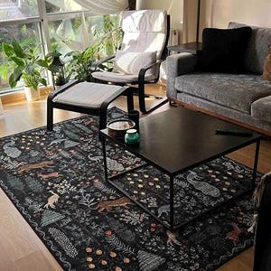 May include: A black area rug with a detailed forest pattern featuring animals and foliage. The rug is placed in a living room setting with a black coffee table, a chair, and a sofa. The room has large windows and plants.