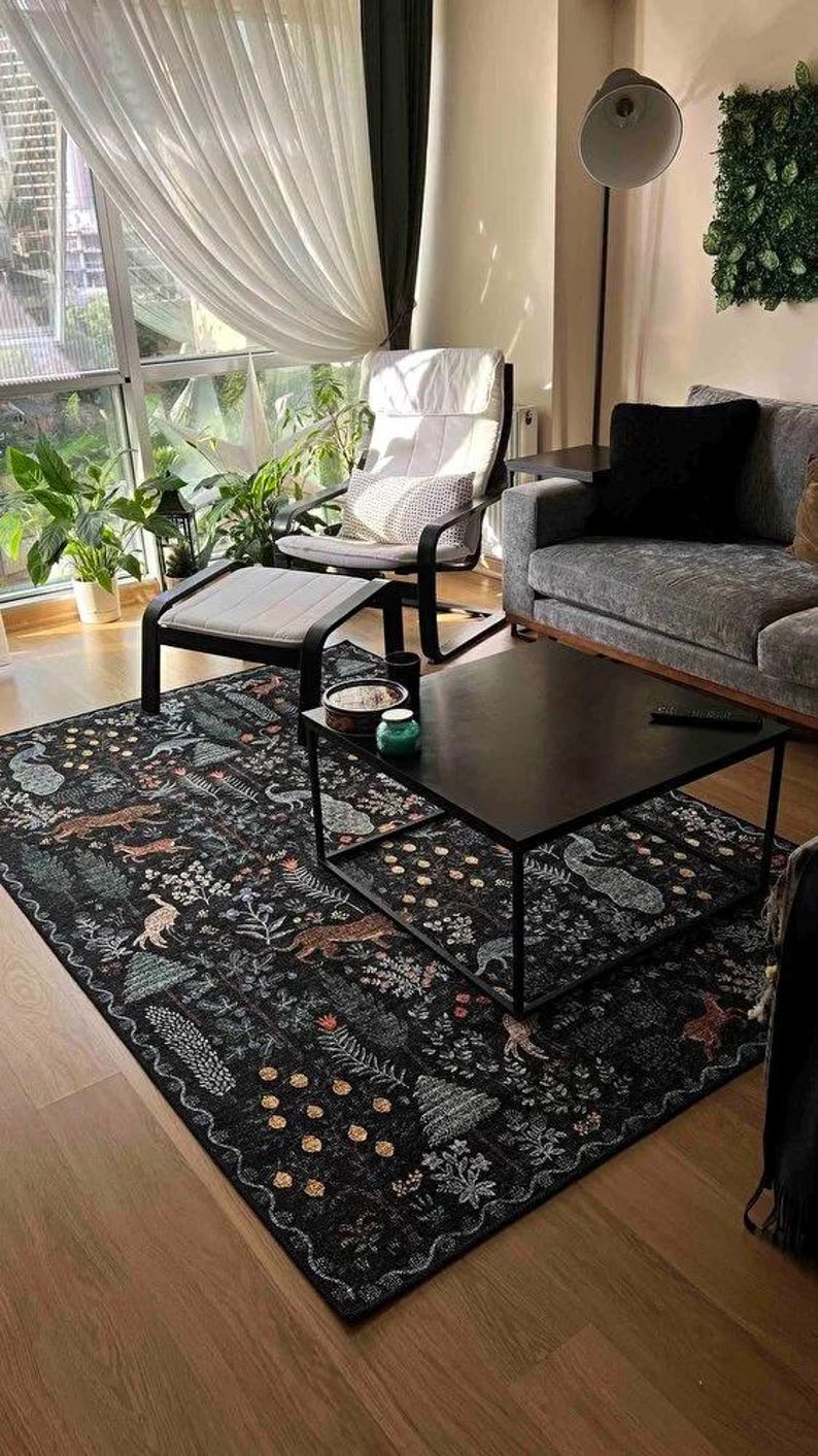 May include: A black area rug with a detailed forest pattern featuring animals and foliage. The rug is placed in a living room setting with a black coffee table, a chair, and a sofa. The room has large windows and plants.