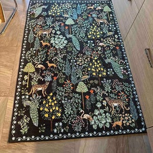 May include: A rectangular black rug with a detailed forest scene. The rug features trees, flowers, deer, leopards, flamingos, and peacocks in shades of green, yellow, orange, and white. The border has a white, wavy pattern.