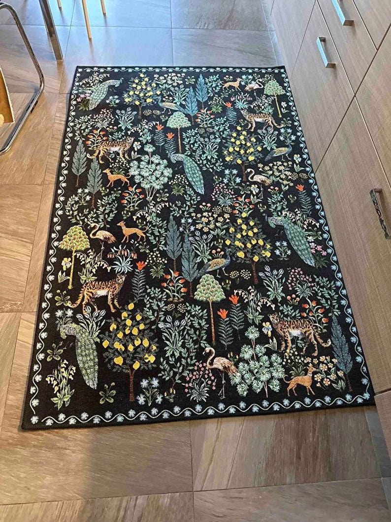 May include: A rectangular black rug with a detailed forest scene. The rug features trees, flowers, deer, leopards, flamingos, and peacocks in shades of green, yellow, orange, and white. The border has a white, wavy pattern.
