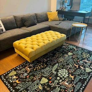 May include: A living room scene featuring a large gray sectional sofa, a yellow tufted ottoman, and a black rug with a forest design. The rug has a detailed pattern of trees, plants, and animals. A small side table and a desk are also visible.