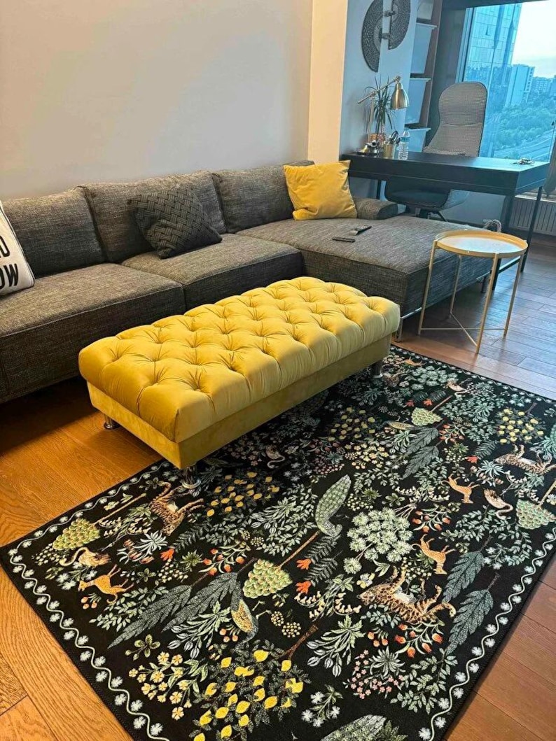 May include: A living room scene featuring a large gray sectional sofa, a yellow tufted ottoman, and a black rug with a forest design. The rug has a detailed pattern of trees, plants, and animals. A small side table and a desk are also visible.
