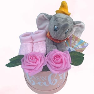 May include: A baby gift basket with a grey Dumbo the Elephant soft toy, pink baby socks, and two pink roses. The arrangement is in a round, pale pink box with the words "NEW BABY" in white script.