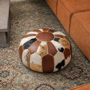 Peut inclure: Pouf rond en cuir avec un motif patchwork. Le pouf présente un mélange de pièces en peau de vache marron, blanche et noire. Le dessus est un cercle en cuir marron uni, et le design est géométrique.