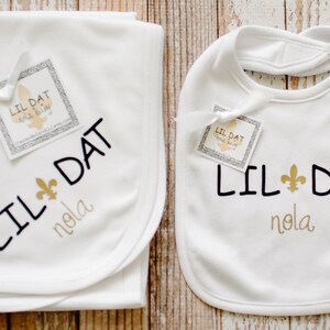 May include: White baby blanket and bib with black embroidery that says "Lil Dat Nola" and a gold fleur-de-lis.