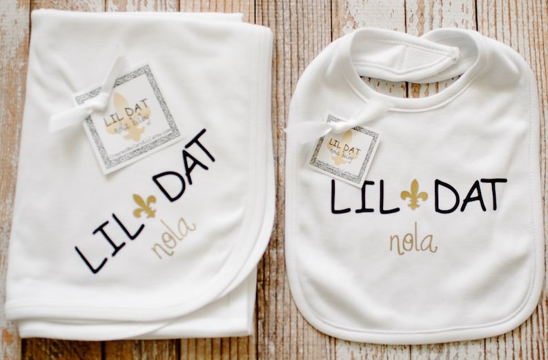May include: White baby blanket and bib with black embroidery that says "Lil Dat Nola" and a gold fleur-de-lis.