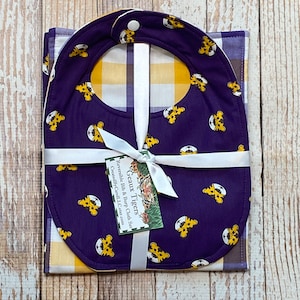 May include: A reversible bib and burp cloth set. The bib is dark purple with a pattern of yellow tiger faces and soccer balls. The burp cloth has a yellow and white plaid pattern. The set is tied with a white ribbon and a tag that reads "Geaux Tigers".