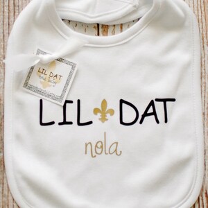 May include: White baby bib with black text that reads "Lil Dat" with a gold fleur-de-lis symbol and "nola" in gold script.