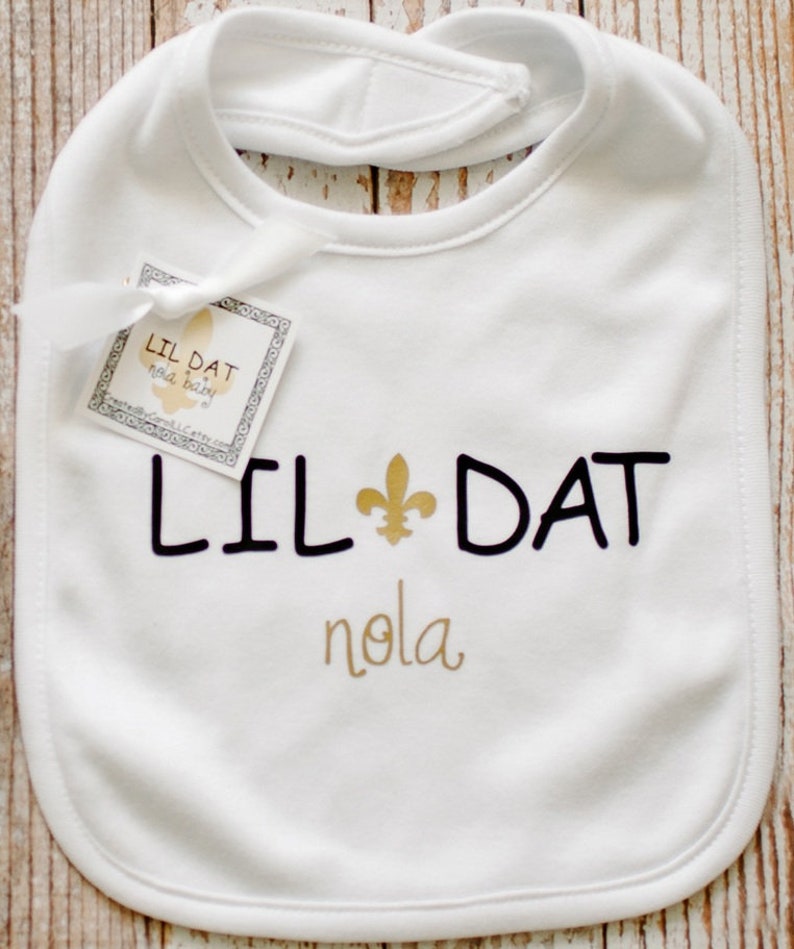 May include: White baby bib with black text that reads "Lil Dat" with a gold fleur-de-lis symbol and "nola" in gold script.