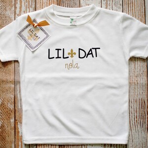 May include: White t-shirt with black text that reads "Lil Dat" with a gold fleur-de-lis and "nola" in gold lettering.